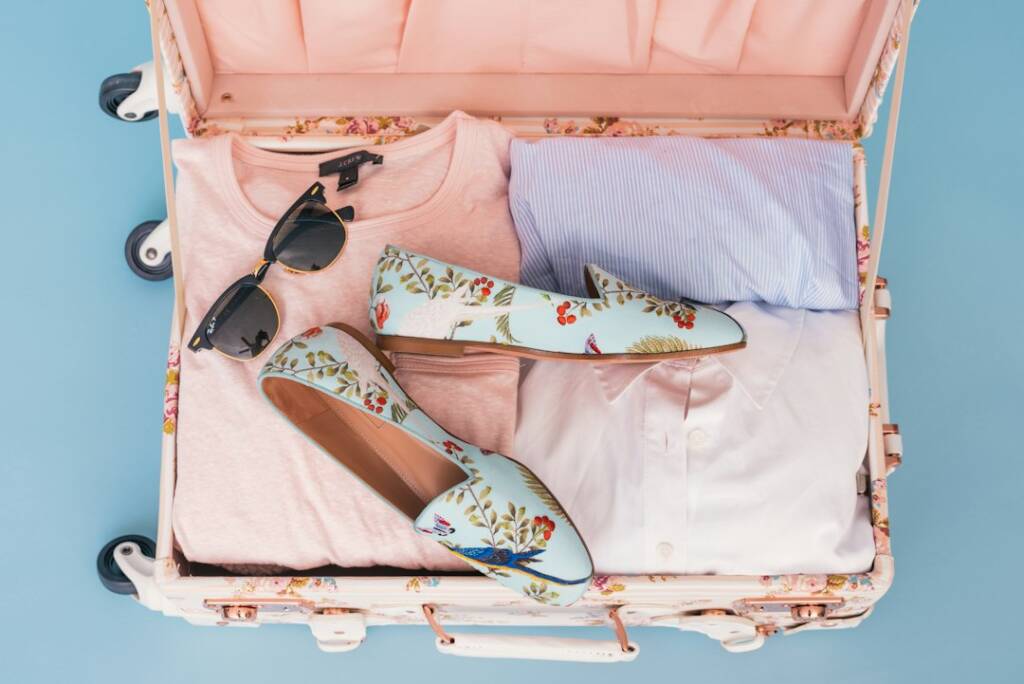 An open pink suitcase packed with folded clothes, sunglasses and floral shoes.